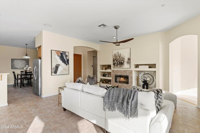 living room with a tiled fireplace, ceiling fan, built in features, and light tile patterned floors