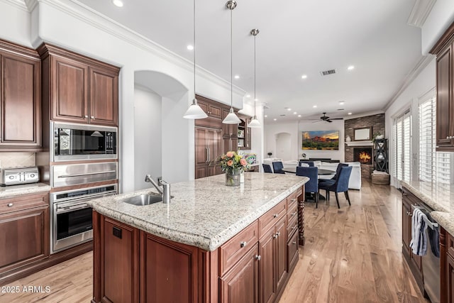 kitchen with a large fireplace, a kitchen island with sink, a sink, stainless steel appliances, and light wood finished floors