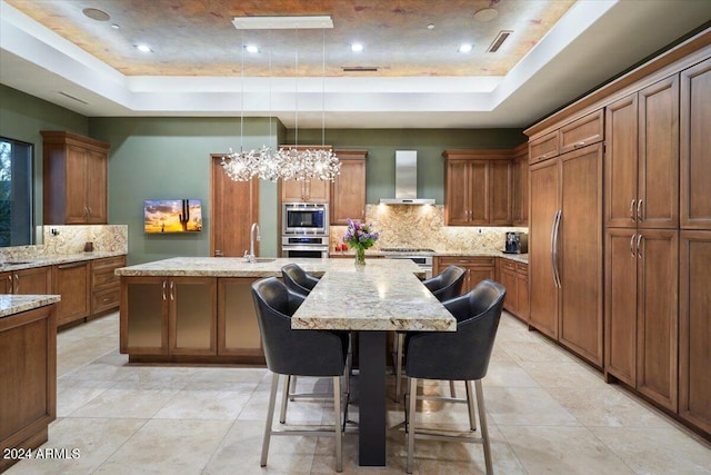 kitchen with a raised ceiling, built in appliances, an island with sink, and wall chimney range hood