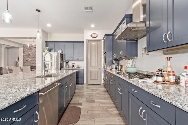 kitchen with pendant lighting, appliances with stainless steel finishes, wall chimney exhaust hood, sink, and light stone counters