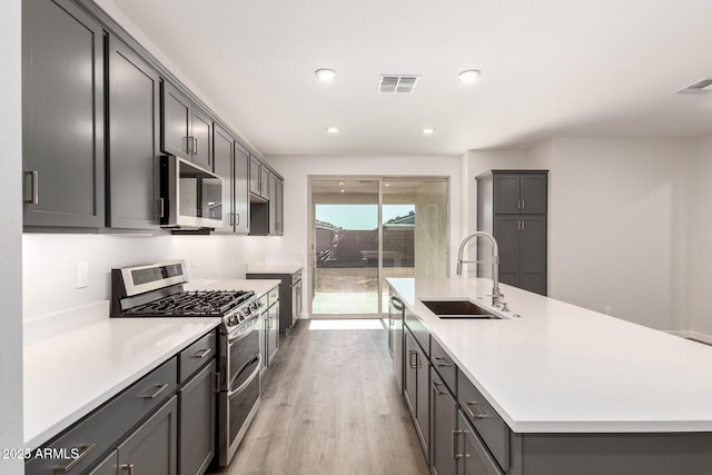 kitchen featuring sink, light wood-type flooring, gray cabinets, a kitchen island with sink, and appliances with stainless steel finishes