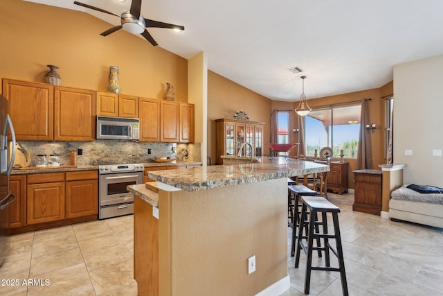 kitchen with light stone counters, hanging light fixtures, appliances with stainless steel finishes, an island with sink, and a kitchen bar