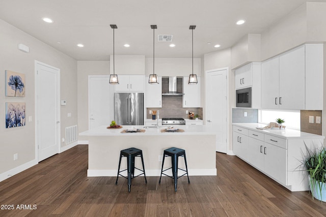 kitchen with white cabinetry, high quality fridge, wall chimney exhaust hood, hanging light fixtures, and a kitchen island with sink