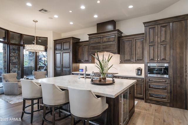 kitchen with pendant lighting, light hardwood / wood-style flooring, a kitchen island with sink, dark brown cabinets, and stainless steel oven