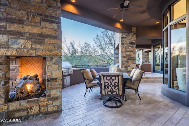 patio terrace at dusk featuring area for grilling, ceiling fan, and an outdoor stone fireplace