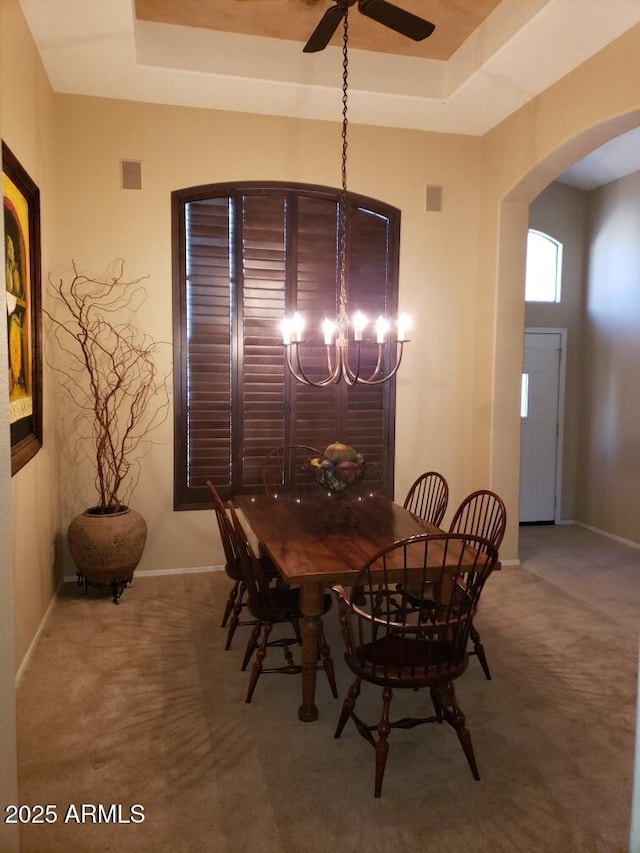 carpeted dining space with ceiling fan with notable chandelier and a tray ceiling