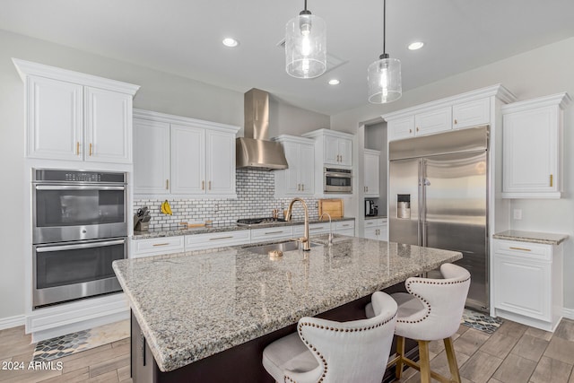 kitchen with wall chimney exhaust hood, stainless steel appliances, a center island with sink, tasteful backsplash, and pendant lighting