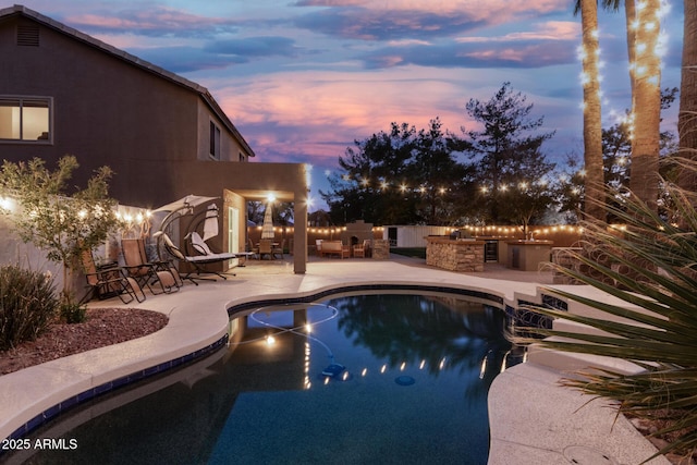 pool at dusk featuring an outdoor kitchen, a fireplace, and a patio area