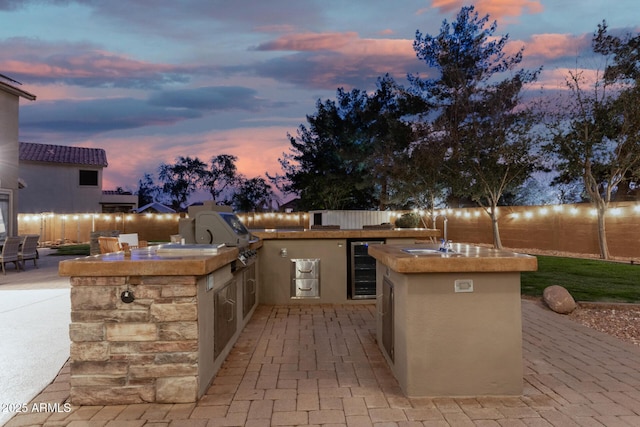 patio terrace at dusk with an outdoor kitchen, sink, beverage cooler, and area for grilling