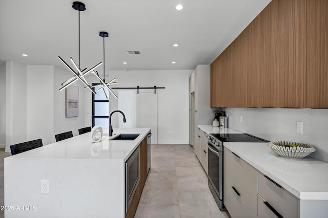 kitchen with a barn door, stainless steel appliances, a kitchen island with sink, hanging light fixtures, and white cabinets