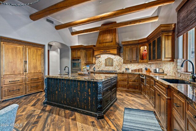 kitchen with beam ceiling, sink, dark wood-type flooring, light stone counters, and an island with sink