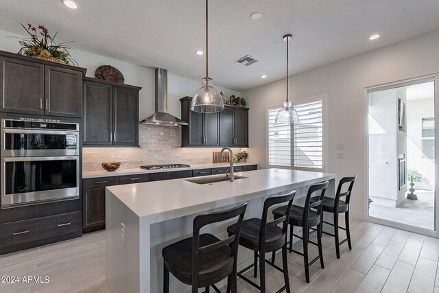 kitchen featuring appliances with stainless steel finishes, an island with sink, wall chimney exhaust hood, decorative light fixtures, and sink