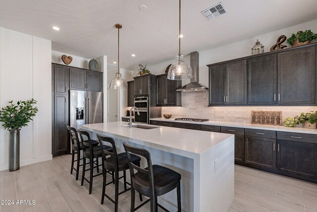 kitchen featuring hanging light fixtures, stainless steel appliances, a kitchen island with sink, sink, and wall chimney range hood