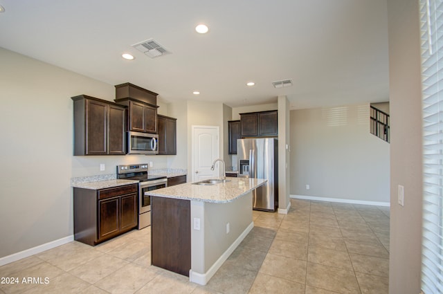 kitchen with a kitchen island with sink, sink, stainless steel appliances, dark brown cabinetry, and light stone countertops