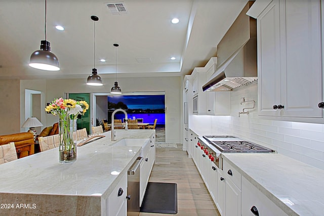 kitchen featuring decorative light fixtures, wall chimney range hood, white cabinets, a kitchen island with sink, and light stone counters