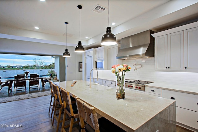 kitchen featuring a center island with sink, wall chimney exhaust hood, white cabinetry, a water view, and sink