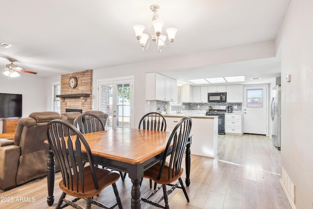 dining area with a stone fireplace, light wood-style flooring, visible vents, and ceiling fan with notable chandelier