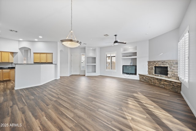 unfurnished living room featuring a stone fireplace, dark wood-type flooring, ceiling fan, and built in shelves