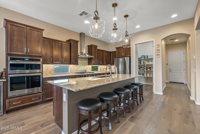 kitchen with wall chimney range hood, sink, hanging light fixtures, an island with sink, and stainless steel appliances