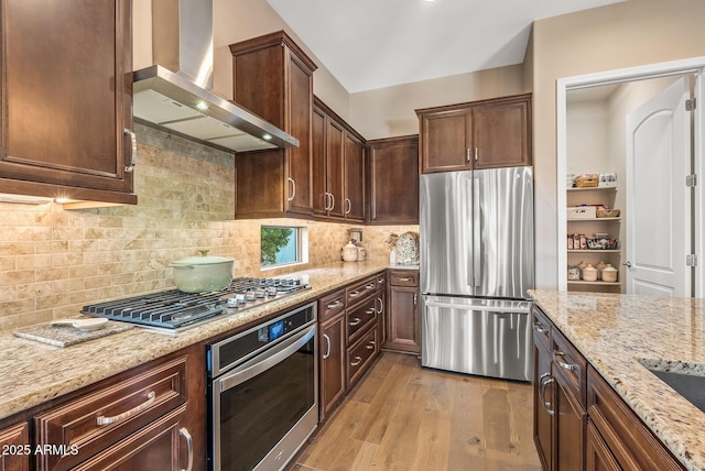 kitchen with wall chimney exhaust hood, light stone counters, backsplash, light hardwood / wood-style floors, and appliances with stainless steel finishes