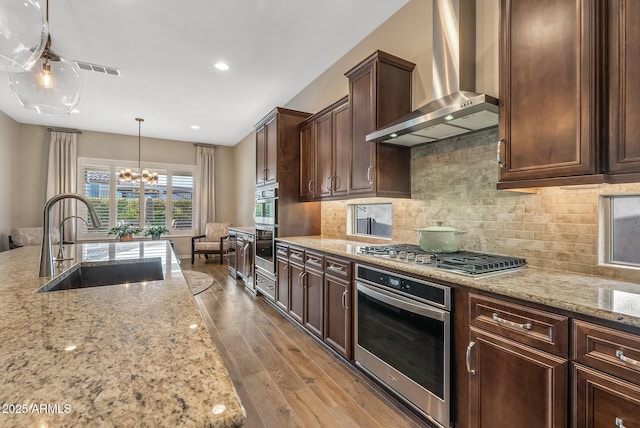 kitchen with light stone counters, stainless steel appliances, sink, wall chimney range hood, and a chandelier