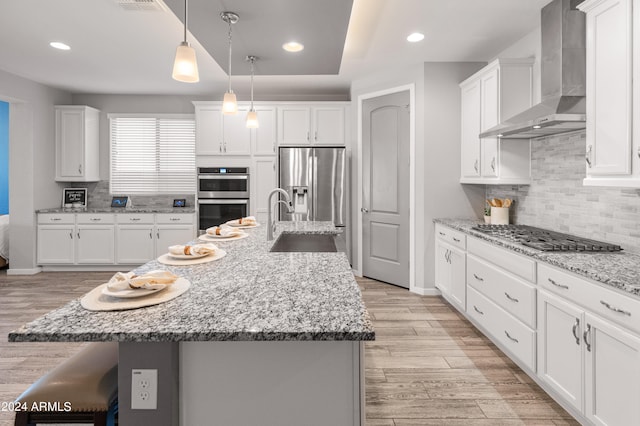 kitchen featuring appliances with stainless steel finishes, an island with sink, wall chimney range hood, and hanging light fixtures