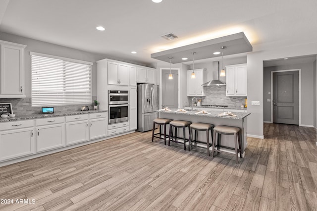 kitchen featuring white cabinets, appliances with stainless steel finishes, light wood-type flooring, wall chimney exhaust hood, and light stone counters