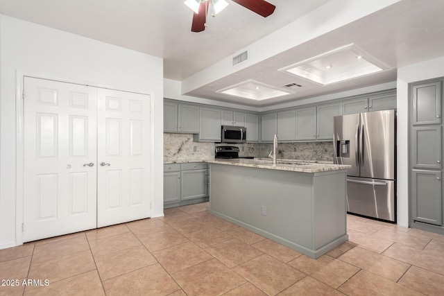 kitchen featuring light tile patterned flooring, gray cabinetry, appliances with stainless steel finishes, a raised ceiling, and a kitchen island with sink