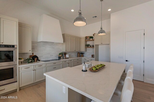 kitchen with black electric cooktop, custom range hood, decorative backsplash, double oven, and light hardwood / wood-style flooring