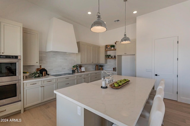 kitchen featuring an island with sink, black electric stovetop, light stone counters, custom range hood, and stainless steel double oven