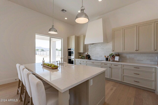 kitchen featuring tasteful backsplash, a center island with sink, double oven, light hardwood / wood-style floors, and wall chimney exhaust hood