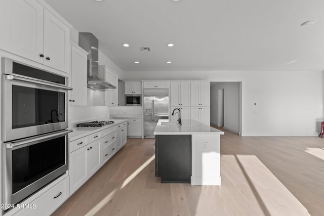 kitchen featuring wall chimney range hood, built in appliances, light hardwood / wood-style floors, white cabinets, and a center island with sink