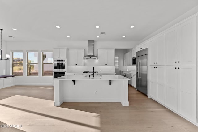 kitchen featuring a breakfast bar area, white cabinets, a kitchen island with sink, stainless steel appliances, and wall chimney range hood