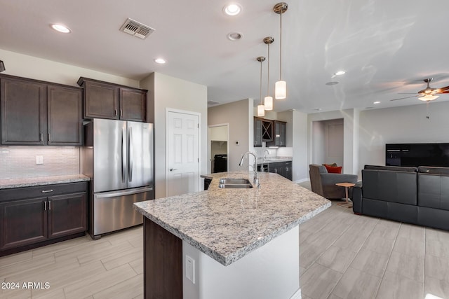 kitchen featuring dark brown cabinetry, sink, stainless steel fridge, an island with sink, and decorative light fixtures