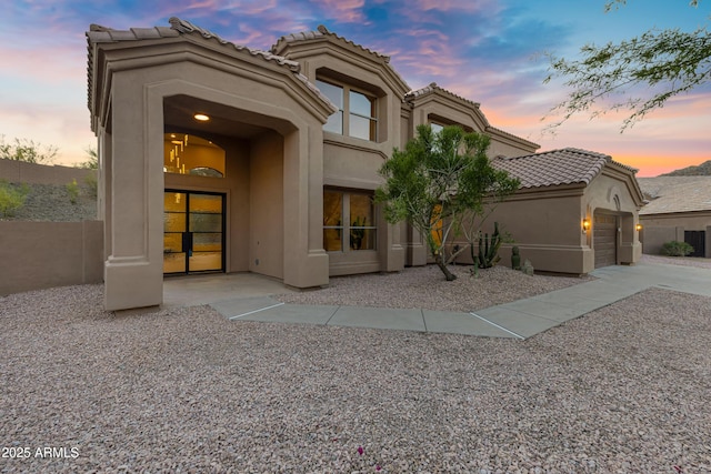 back of house at dusk with an attached garage, a tiled roof, and stucco siding