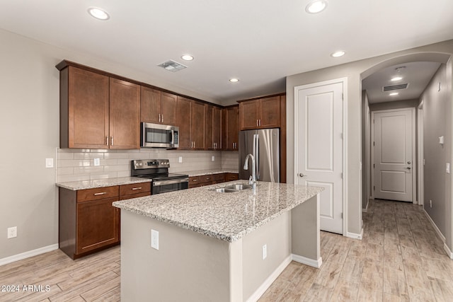 kitchen featuring sink, stainless steel appliances, light stone counters, an island with sink, and light wood-type flooring