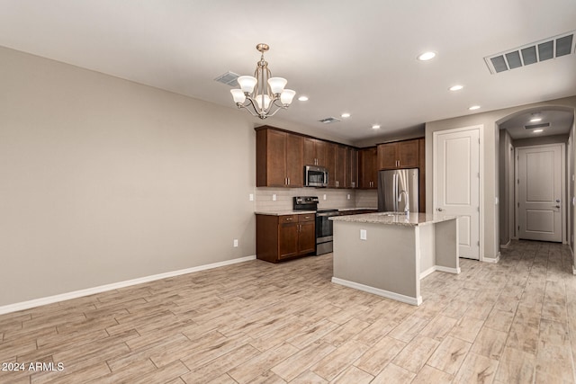 kitchen with light stone countertops, decorative light fixtures, a kitchen island with sink, appliances with stainless steel finishes, and light wood-type flooring