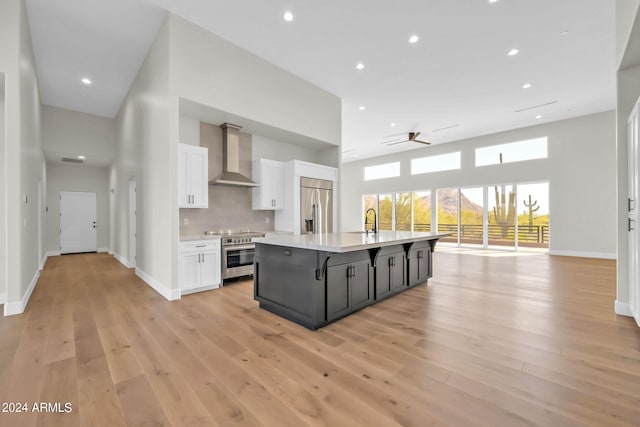 kitchen featuring white cabinets, an island with sink, wall chimney exhaust hood, light hardwood / wood-style flooring, and appliances with stainless steel finishes