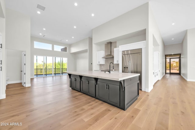 kitchen with white cabinets, built in refrigerator, wall chimney exhaust hood, a towering ceiling, and a center island with sink