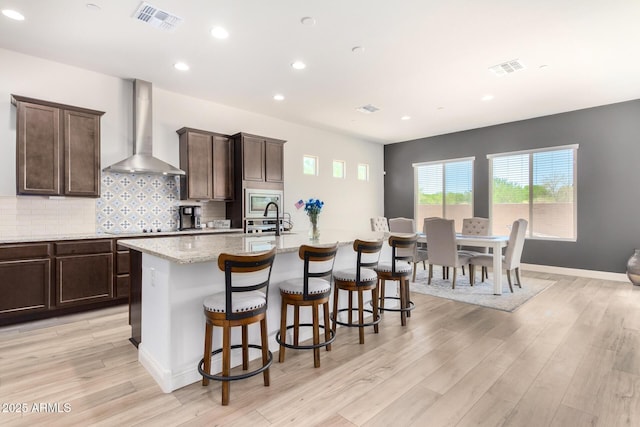 kitchen with visible vents, light wood-style flooring, a center island with sink, tasteful backsplash, and wall chimney range hood