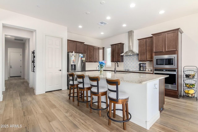 kitchen with visible vents, a kitchen island with sink, stainless steel appliances, wall chimney exhaust hood, and light wood-type flooring