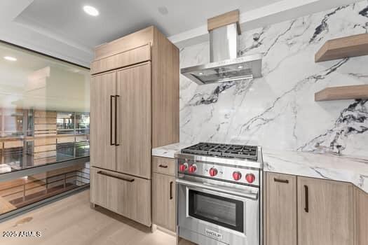 kitchen with tasteful backsplash, light brown cabinetry, light stone counters, premium appliances, and wall chimney exhaust hood