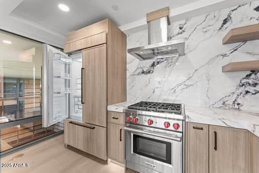 kitchen with tasteful backsplash, light stone countertops, light brown cabinetry, premium stove, and wall chimney exhaust hood