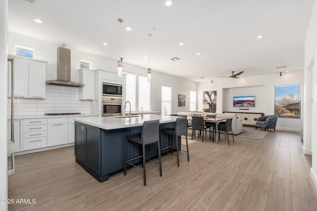 kitchen with pendant lighting, wall chimney range hood, a kitchen island with sink, stainless steel appliances, and white cabinets