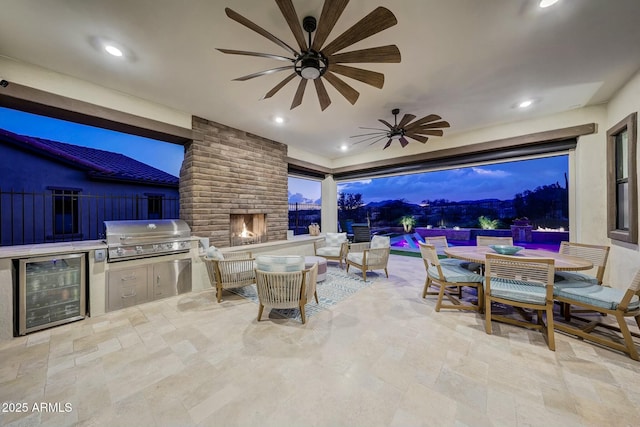 patio terrace at dusk with an outdoor brick fireplace, a swimming pool, wine cooler, area for grilling, and exterior kitchen