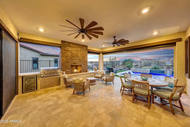 patio terrace at dusk with ceiling fan, an outdoor living space with a fireplace, a grill, area for grilling, and beverage cooler