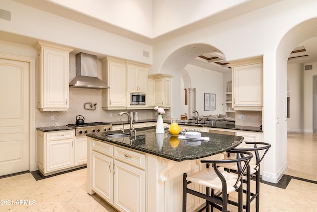 kitchen featuring sink, stainless steel microwave, an island with sink, dark stone counters, and wall chimney exhaust hood