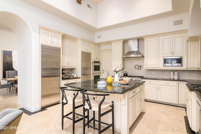 kitchen featuring cream cabinets, a kitchen island with sink, wall chimney range hood, and built in appliances