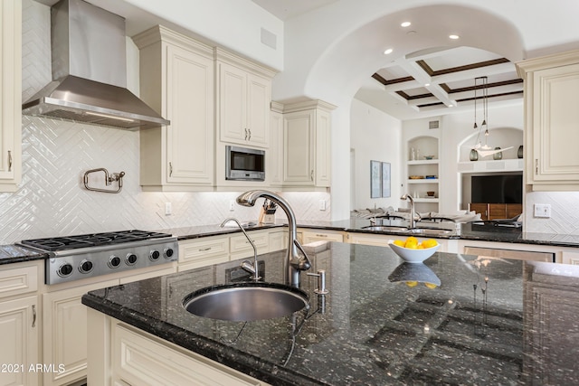 kitchen with sink, dark stone counters, coffered ceiling, cream cabinets, and wall chimney range hood