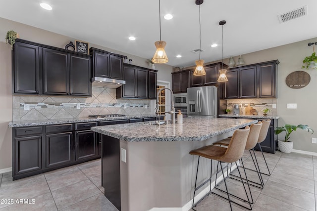kitchen featuring appliances with stainless steel finishes, recessed lighting, light stone counters, an island with sink, and tasteful backsplash